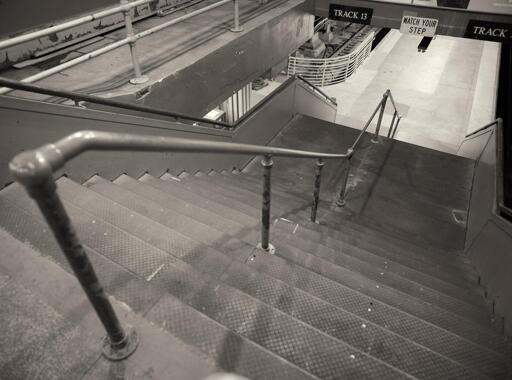 A staircase, with a simple pipe handrail at center, descending to a train platform. Signs above read "Track 13" and "Watch Your Step".