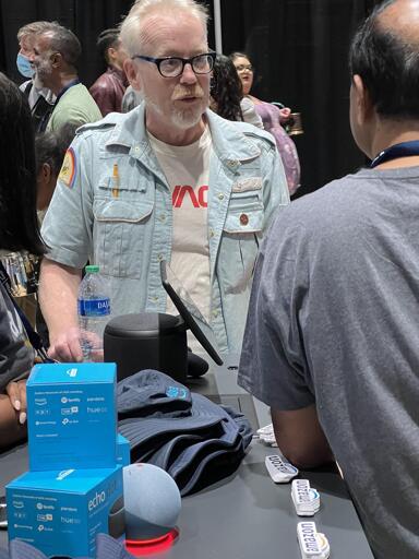 Adam Savage, dressed in a Savage Industries t-shirt and his light blue Nostromo crew shirt, chats with a vendor at booth at SiliCon 2022.