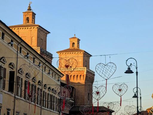 The image captures a charming street scene in Ferrara, Italy, bathed in the warm glow of the late afternoon sun. The brick towers of a historical building rise above, their details highlighted by the sunlight. Hanging across the street are large, heart-shaped festive decorations, adorned with red ribbons and sparkling accents, evoking a romantic, joyful atmosphere. The clear sky contrasts beautifully with the architecture, creating a peaceful, picturesque moment typical of Italian cityscapes.