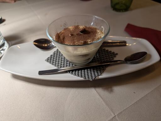 A glass cup filled with tiramisu sits on a white rectangular plate, garnished with cocoa powder and a single coffee bean on top. Two metal spoons are placed on the plate, one resting on a textured gray napkin underneath the dessert. The setting is on a table with a white tablecloth, with a red napkin and a green glass in the background. The lighting casts a soft glow on the dish.