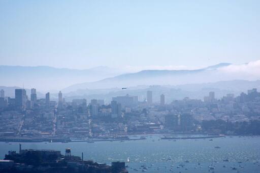 A fighter jet casually flies upside-down and level over San Francisco Bay, with the city (with partial fog) behind it.