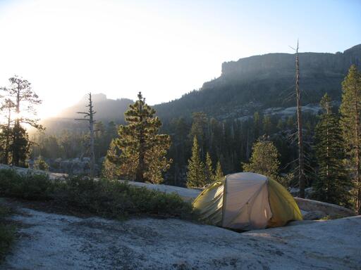 The rising sun washes out this photo in the upper left. Mesa-like mountains are in the background along with many evergreens. In the foreground a small tan and green tent sit at the end of a row of low brush on granite.