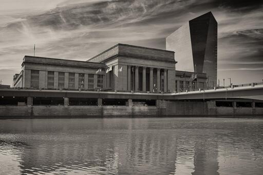 A large neoclassical train station across a river. To the right of the station is a modernist glass office tower.