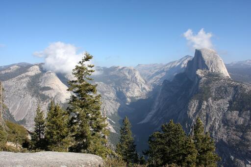 A view looking east, above and into Yosemite Valley, with Half Dome and a pair of cotton-ball clouds. Photo taken from Glacier Point.