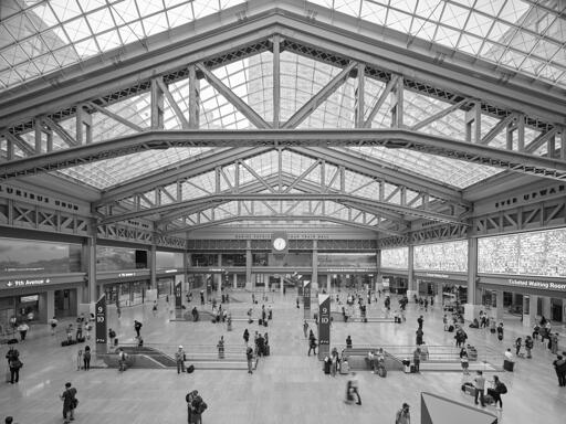 A modern urban passenger rail station hall. Passengers mill about around escalators that descend to unseen tracks below. Exposed steel beams support a skylight roof.