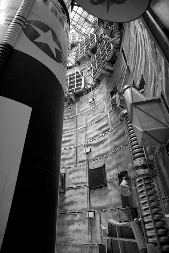 A missile, with a US Air Force logo, pointed up inside a cramped underground silo, which is lined with retractable platforms and various apparatus.
