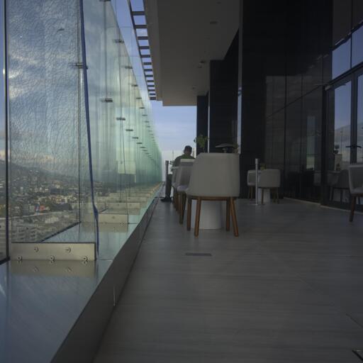 Some weird almost floor to ceiling windows/barriers on a balcony with some two-top tables on it. The leading lines of the wall of glass doors and windows on one side of the balcony & the barriers on the other side point to a distant human figure at the far table. Beyond the barrier to the left you can see a bit of city on a mountain, on the right you can see reflections of sky and a tiny bit of city in the doors & windows. San Jose, Costa Rica. Photo by Peachfront.