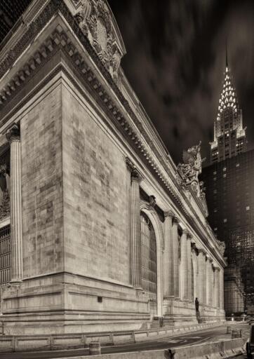 Exterior of a large beaux arts railroad station, with the Chrysler Building in the background, at night. 