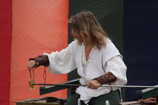 A man with long hair, a loose white Renaissance shirt and a pair of leather arm bracers shapes a hot, hand-blown glass piece.